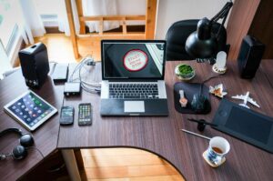 A modern workspace featuring a laptop with START ENGINE STOP on screen, multiple devices, headphones, a coffee cup, and decorative items.
