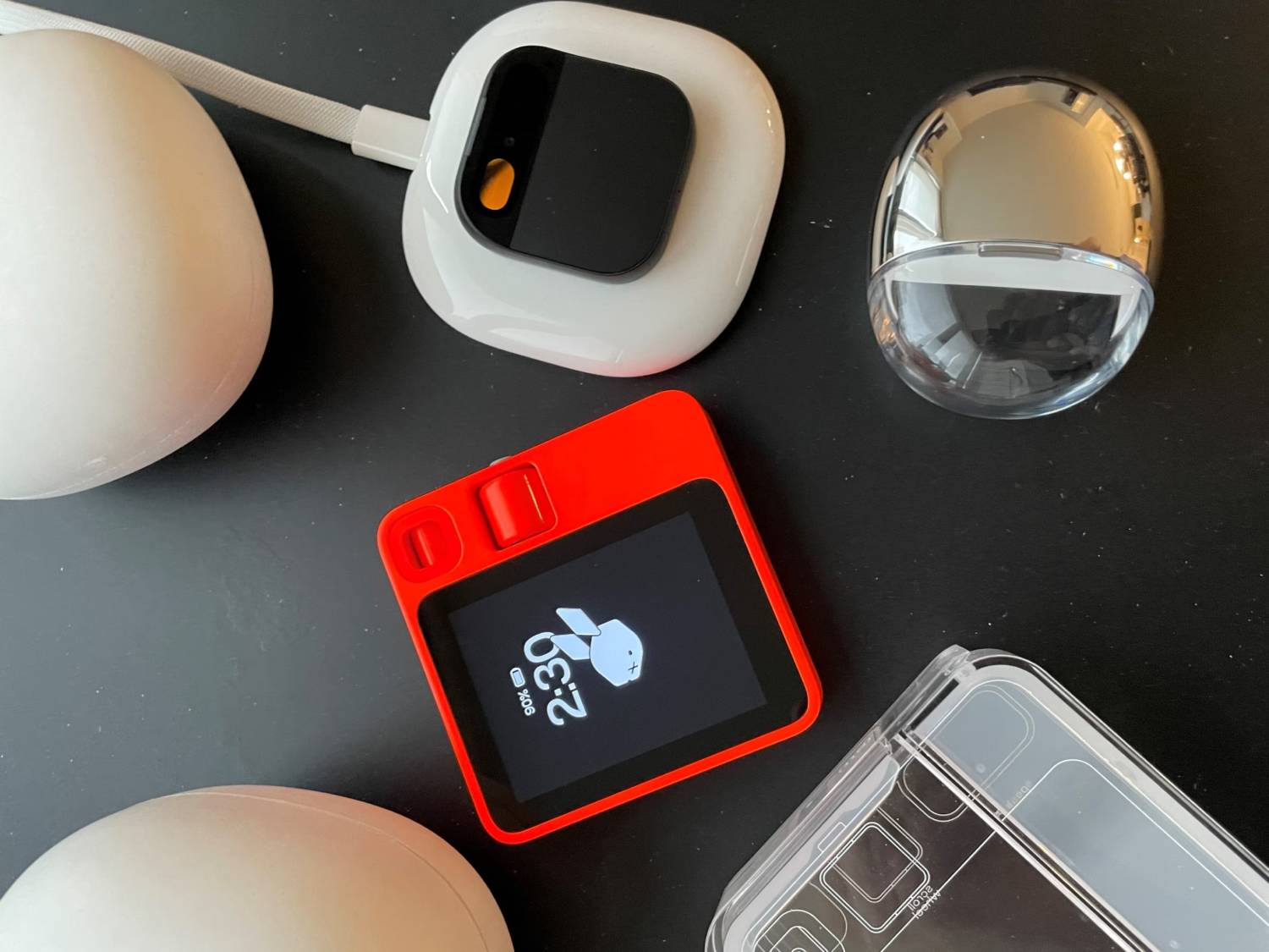 A vibrant red digital device displays the time, surrounded by white earbuds, a charging case, and a sleek, clear cover on a black surface.