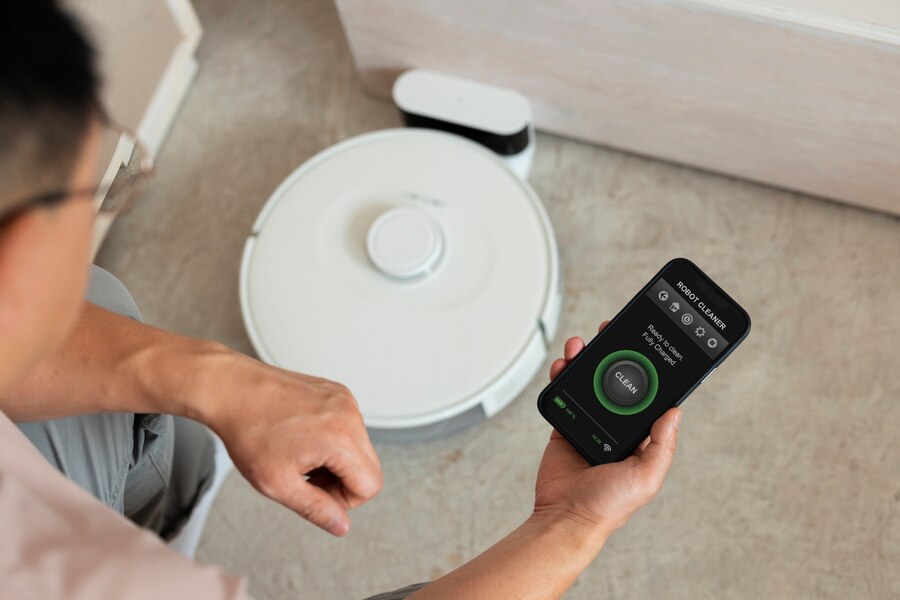 A person using a smartphone to control a robotic vacuum cleaner on a floor, displaying a clean command on the screen.