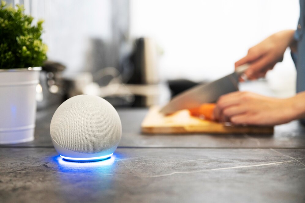 A smart speaker with a blue light rests on a kitchen counter while a person chops vegetables on a wooden cutting board in the background.