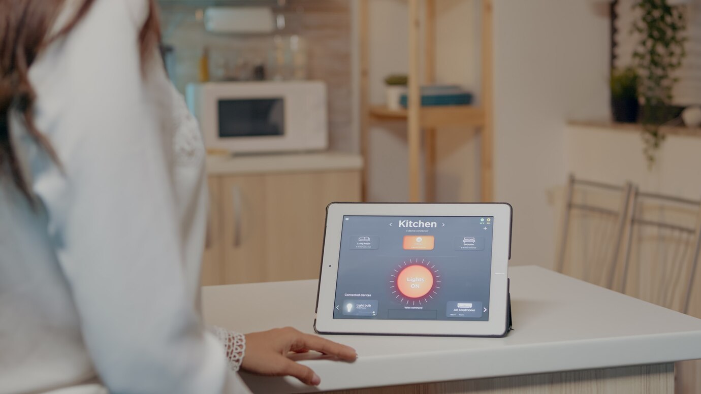 A woman interacts with a tablet displaying smart home controls for the kitchen, including lighting and connected devices.