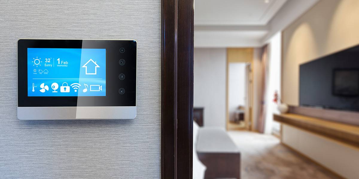 Smart home control panel mounted on a wall, displaying weather, Wi-Fi, and other home automation functions, with a view of a modern living room.