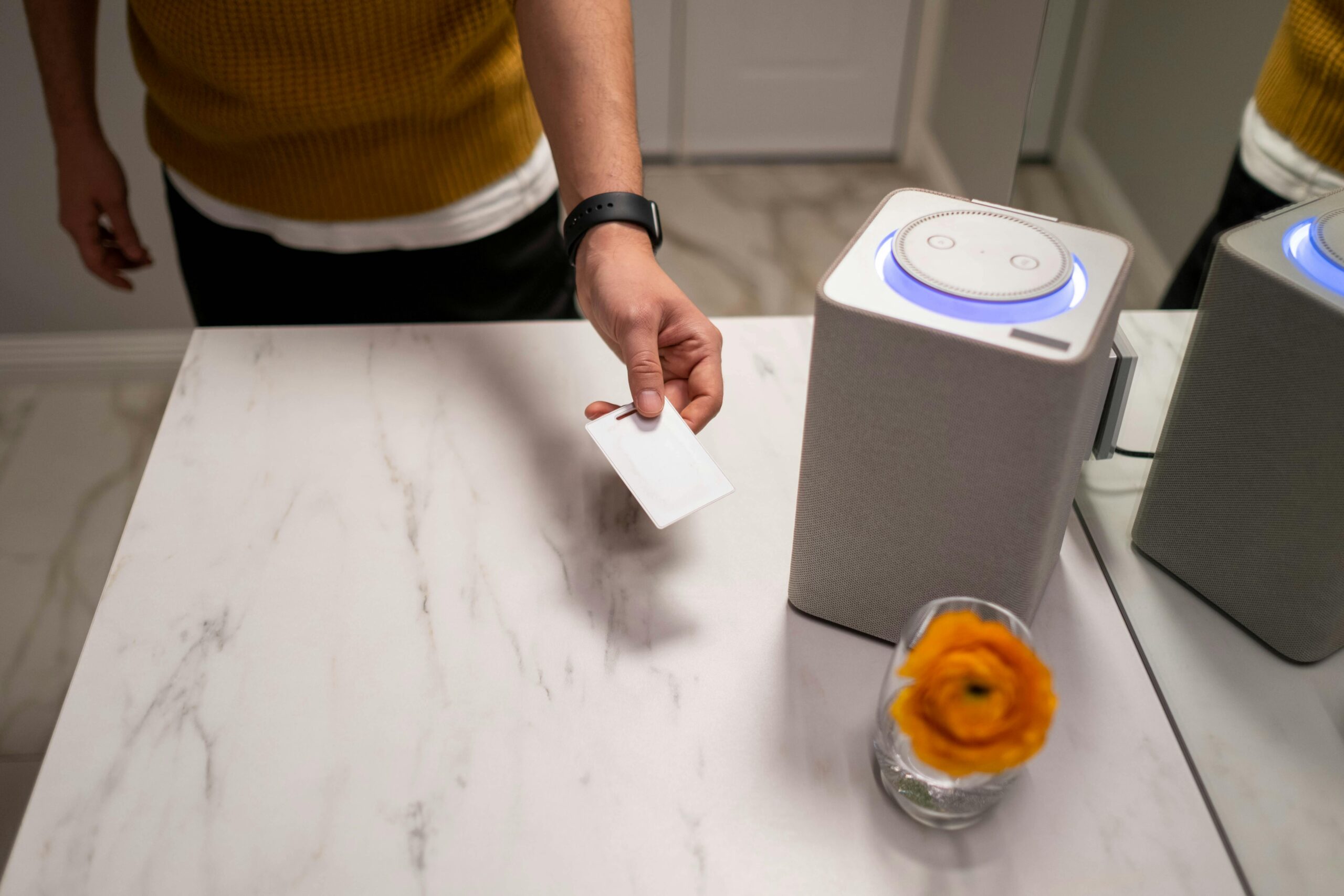 A person in a yellow sweater holds a white card above a stylish speaker on a marble surface, with a flower in a glass nearby.