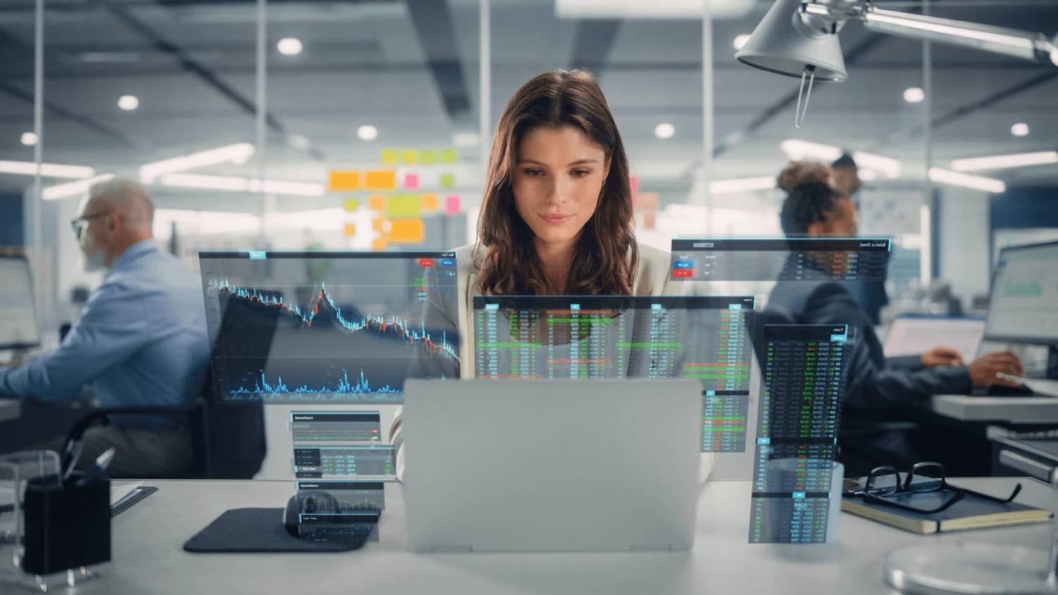 A woman sits at a desk surrounded by multiple screens displaying financial charts and data, in a modern office setting.