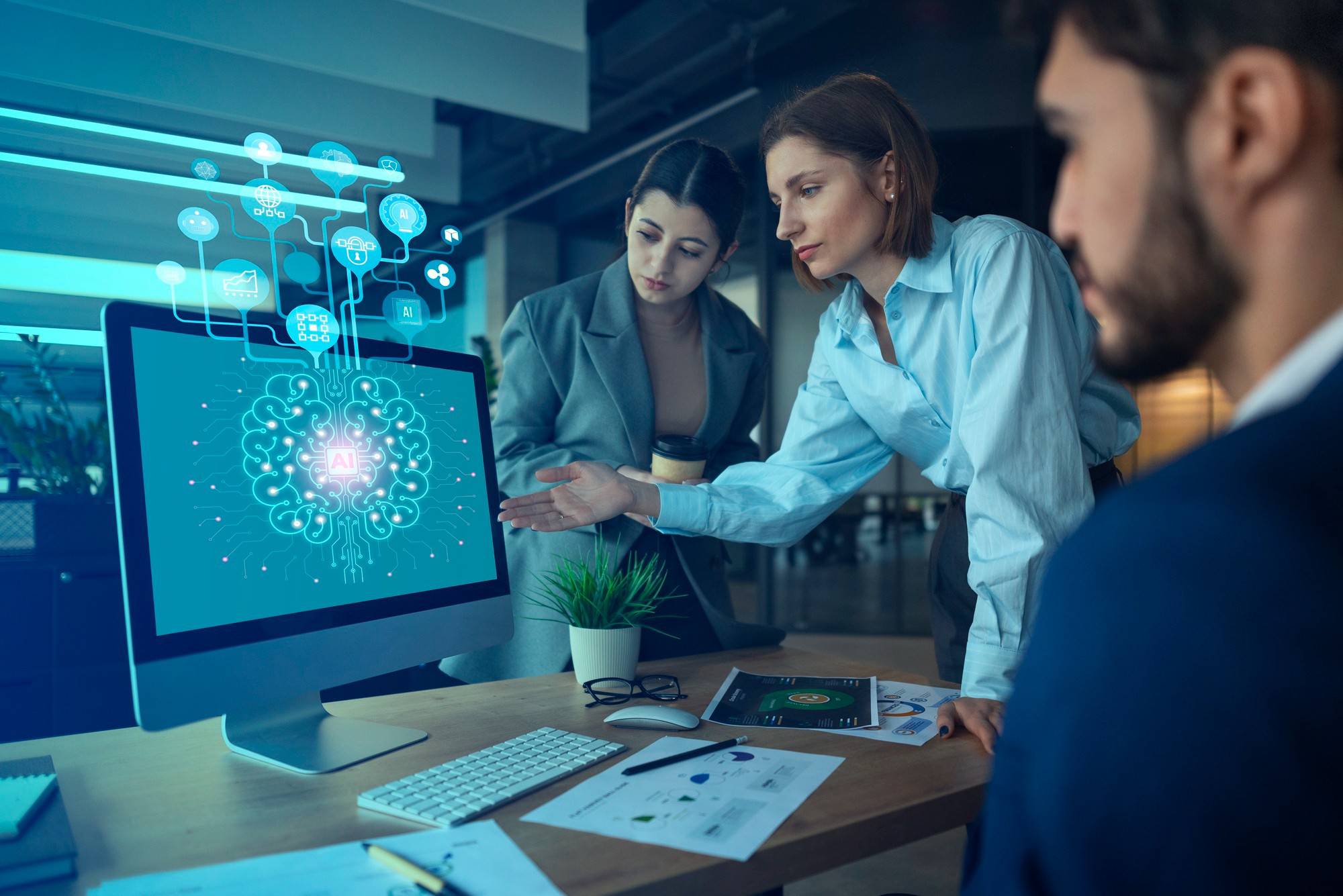 A team collaborates in an office setting, discussing AI concepts displayed on a computer screen surrounded by documents and a plant.