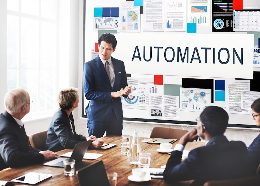 A business meeting with a presenter discussing automation, surrounded by charts and data on a large screen, while attendees look on.