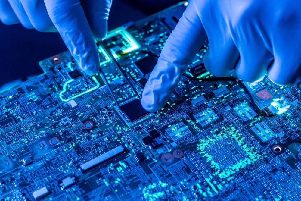 Close-up of a gloved hand delicately working on a glowing blue circuit board, using tweezers to manipulate electronic components.