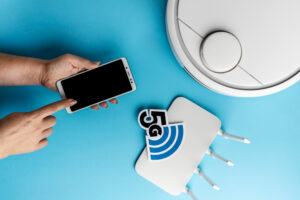 A hand points at a blank smartphone screen, next to a white robotic vacuum and a 5G router on a blue background.