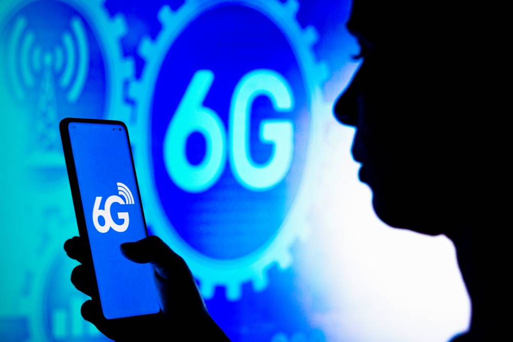  A hand holding a smartphone displays the 6G logo, with a blue background featuring tech-related graphics and symbols.