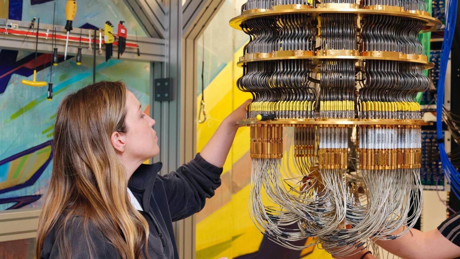  A person interacts with a complex quantum computing device featuring intricate wiring and gold components, set against a colorful background.