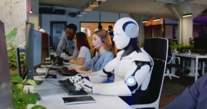 A humanoid robot sits at a desk with computers, surrounded by people working in a modern office filled with plants.