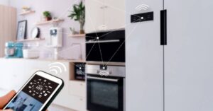 A smartphone displays IoT controls while connected to a modern kitchen with smart appliances, illustrating smart home technology.