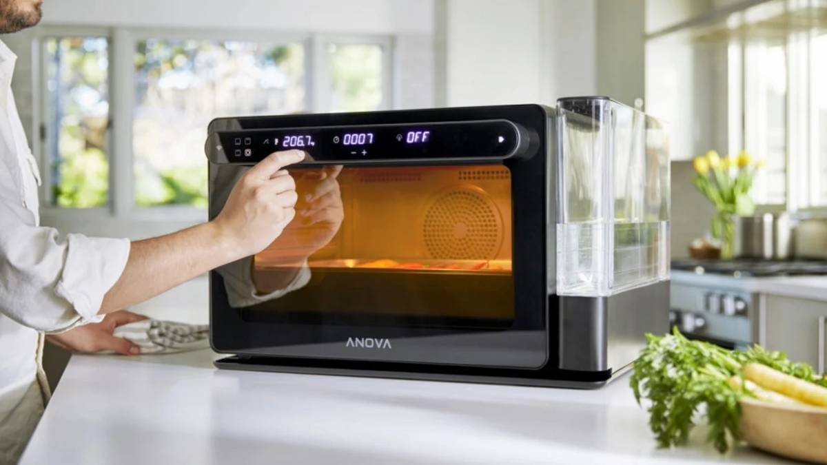 A person adjusts settings on an ANOVA countertop oven with a clear water reservoir, surrounded by a bright kitchen setting.
