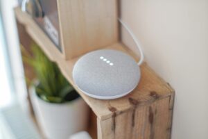 A gray smart speaker on a wooden shelf, with soft glowing lights, beside a green plant in a white pot.