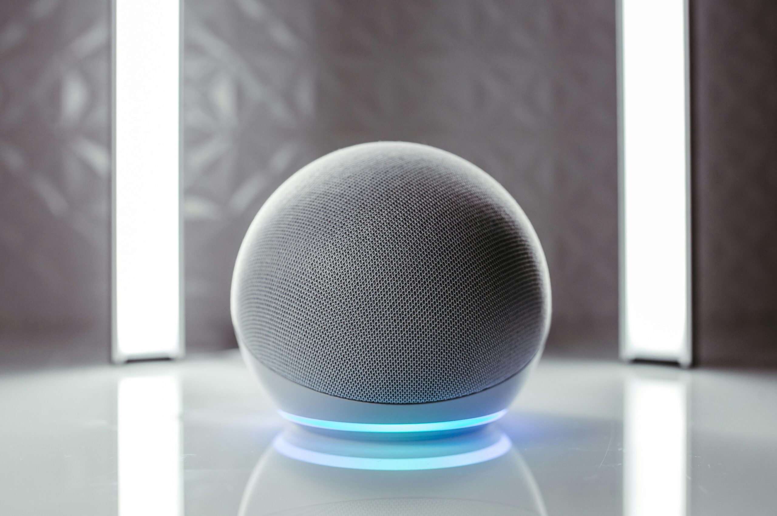 A speaker with a glowing blue base sits on a reflective surface, illuminated by bright vertical lights in the background.