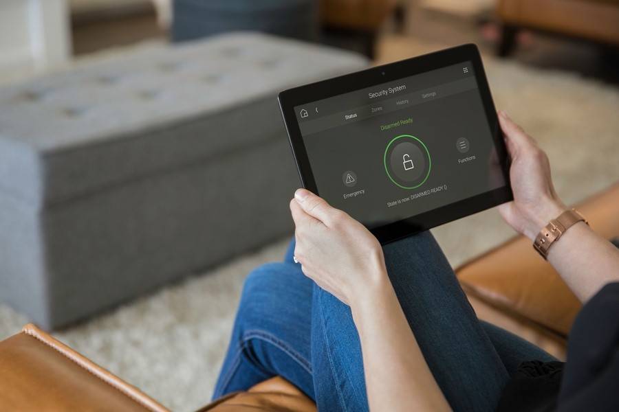 A person holds a tablet displaying a security system interface, indicating Disarmed Ready status while seated in a cozy living room.