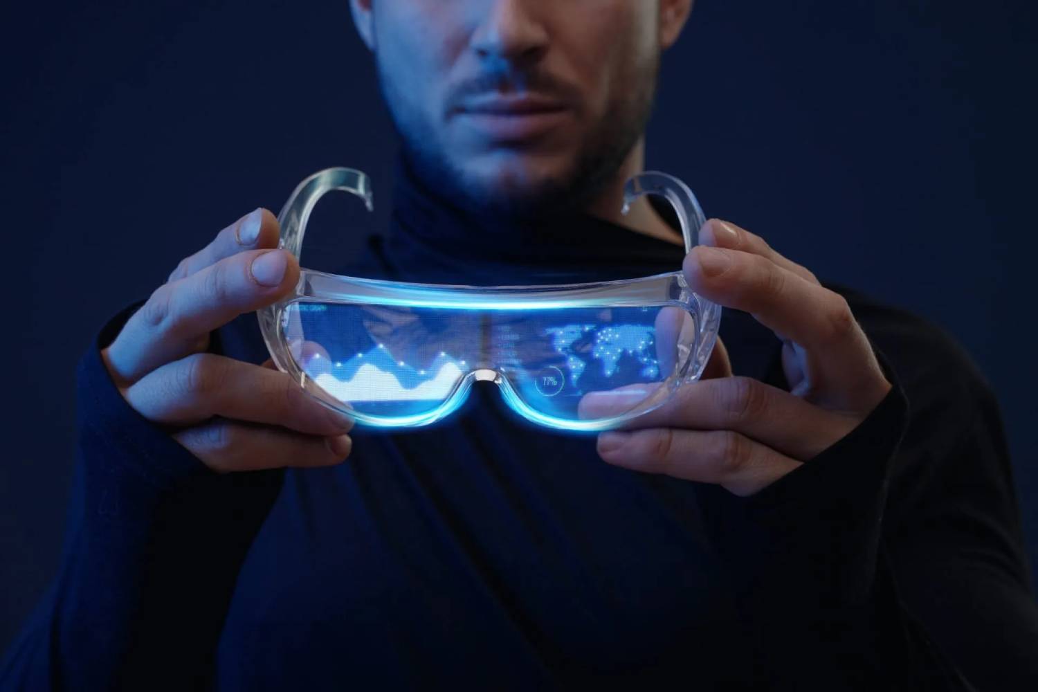  A person holds clear augmented reality glasses displaying blue digital graphics and data against a dark background.