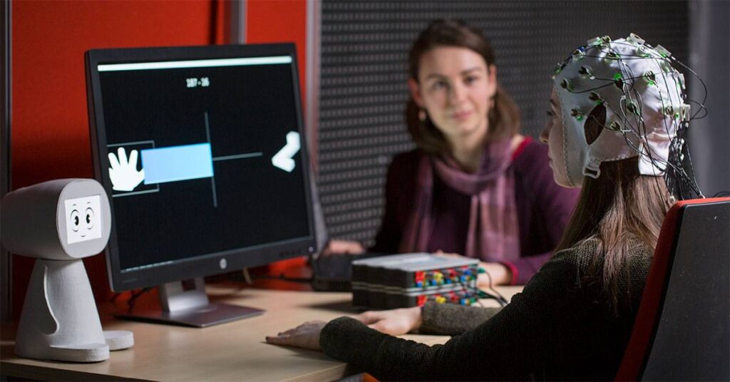 A person wearing an EEG cap interacts with a computer displaying hand graphics, with a robot-like device observing the setup.