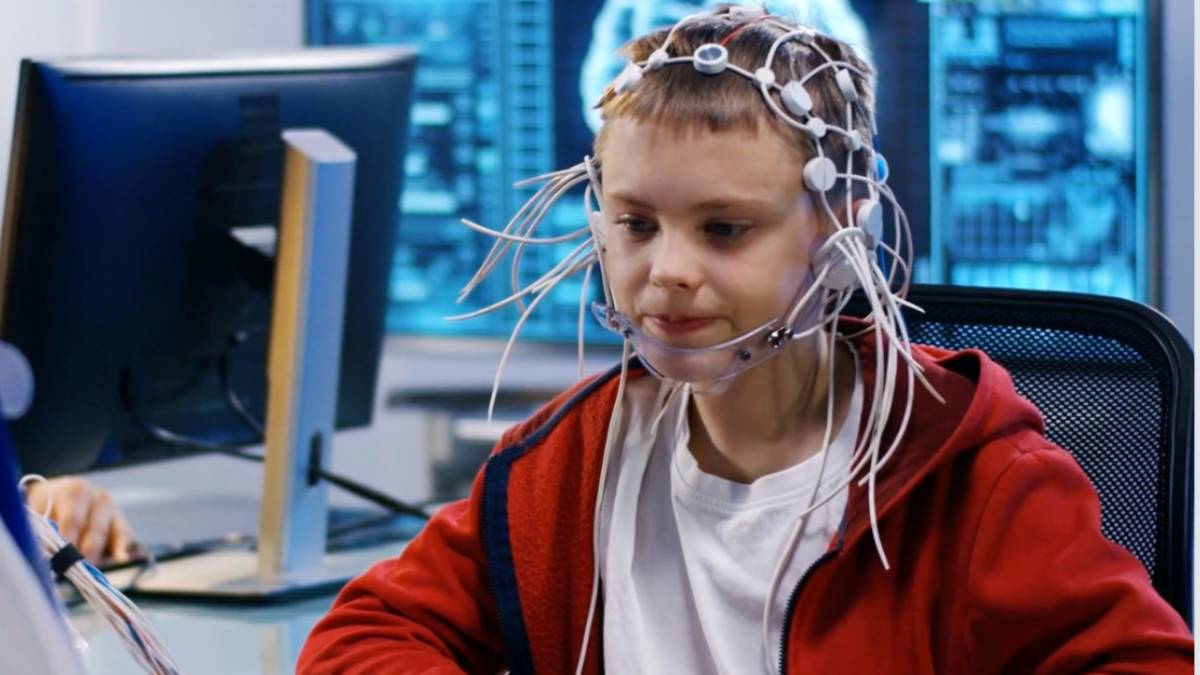  A person wearing a headset with wires sits at a computer in a high-tech environment, indicating a scientific or medical setting.