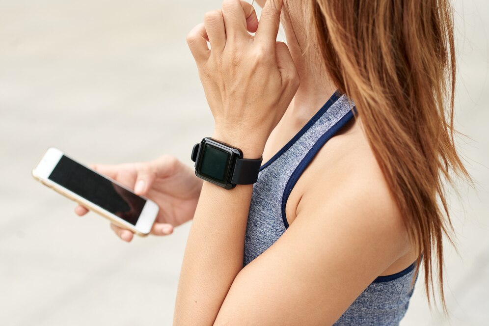  A woman in a sports top checks her smartwatch while holding a smartphone, deep in thought.