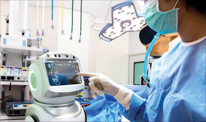 A medical professional in scrubs and gloves interacts with a robotic monitor displaying vital signs in an operating room.