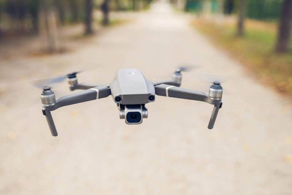 A sleek gray drone with spinning propellers hovers in mid-air above a blurred path lined with trees.