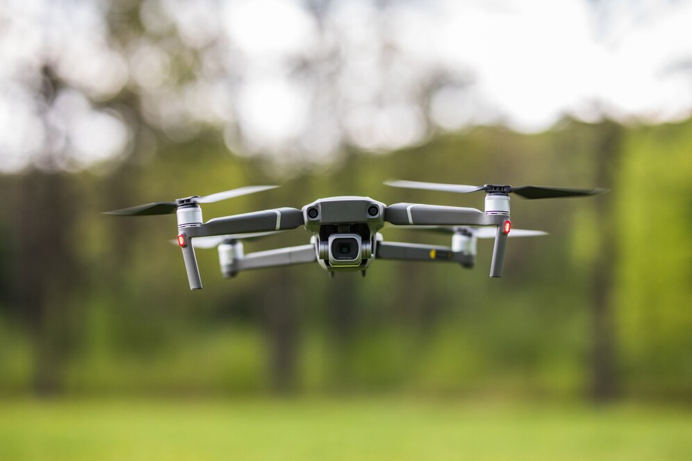A drone with four propellers hovers mid-air against a blurred green background, showcasing its sleek design and camera.