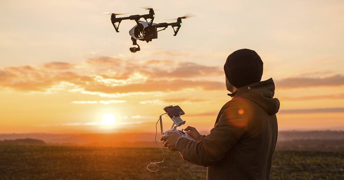 A person controls a drone at sunset, holding a remote with a smartphone, silhouetted against a vibrant sky.