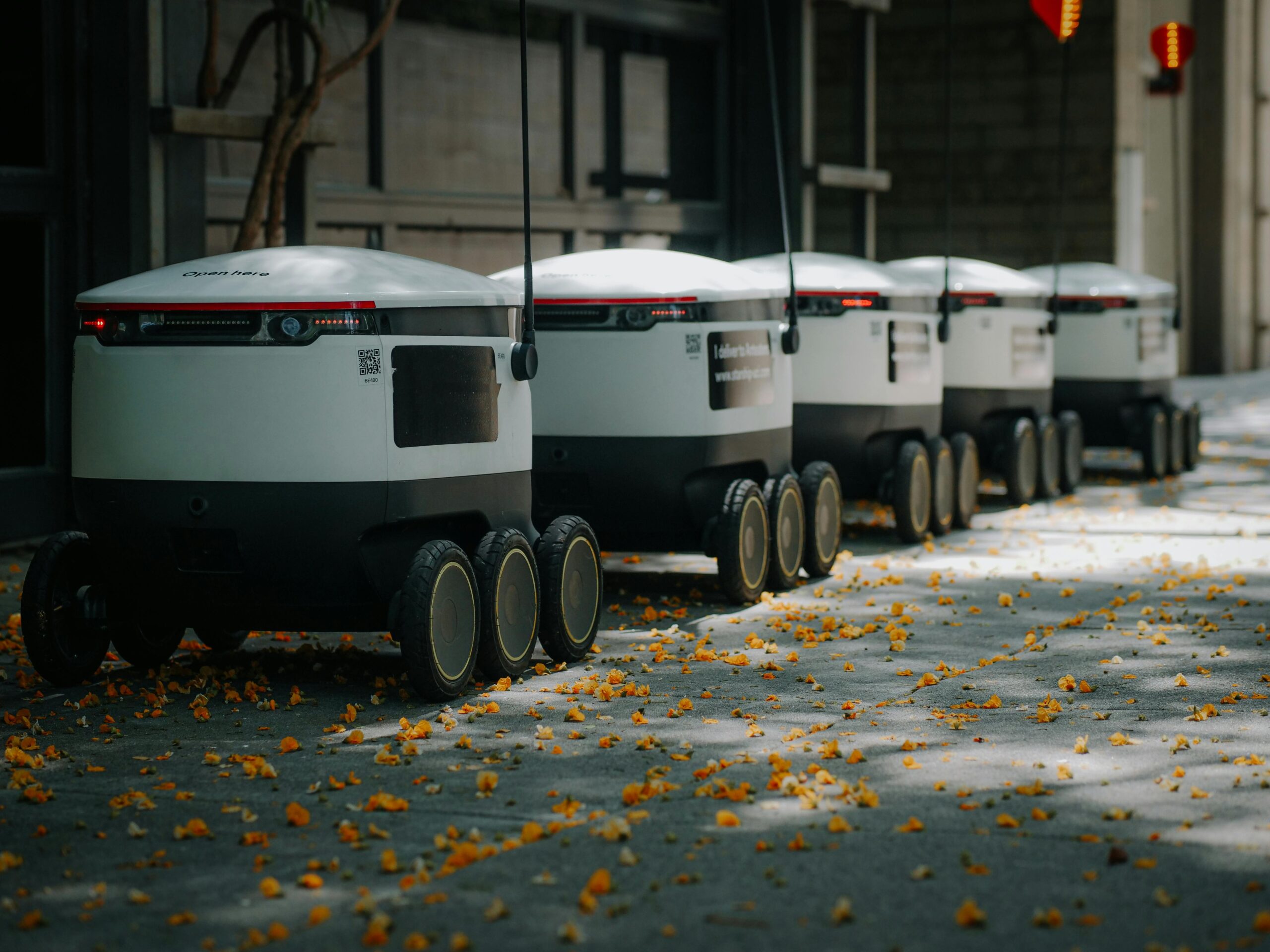 A line of delivery robots on a street covered with orange flower petals, showcasing modern technology in an urban environment.