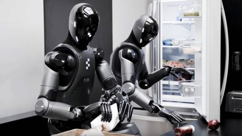 Two humanoid robots engage in a kitchen setting, one preparing food while the other rummages through a refrigerator.