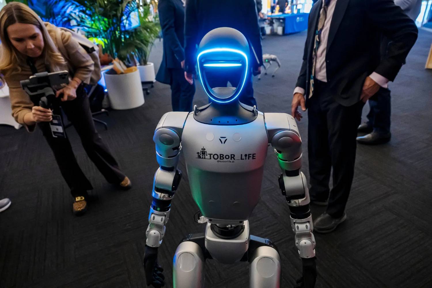 A humanoid robot with glowing blue lights stands in a modern exhibition space, surrounded by people in business attire.