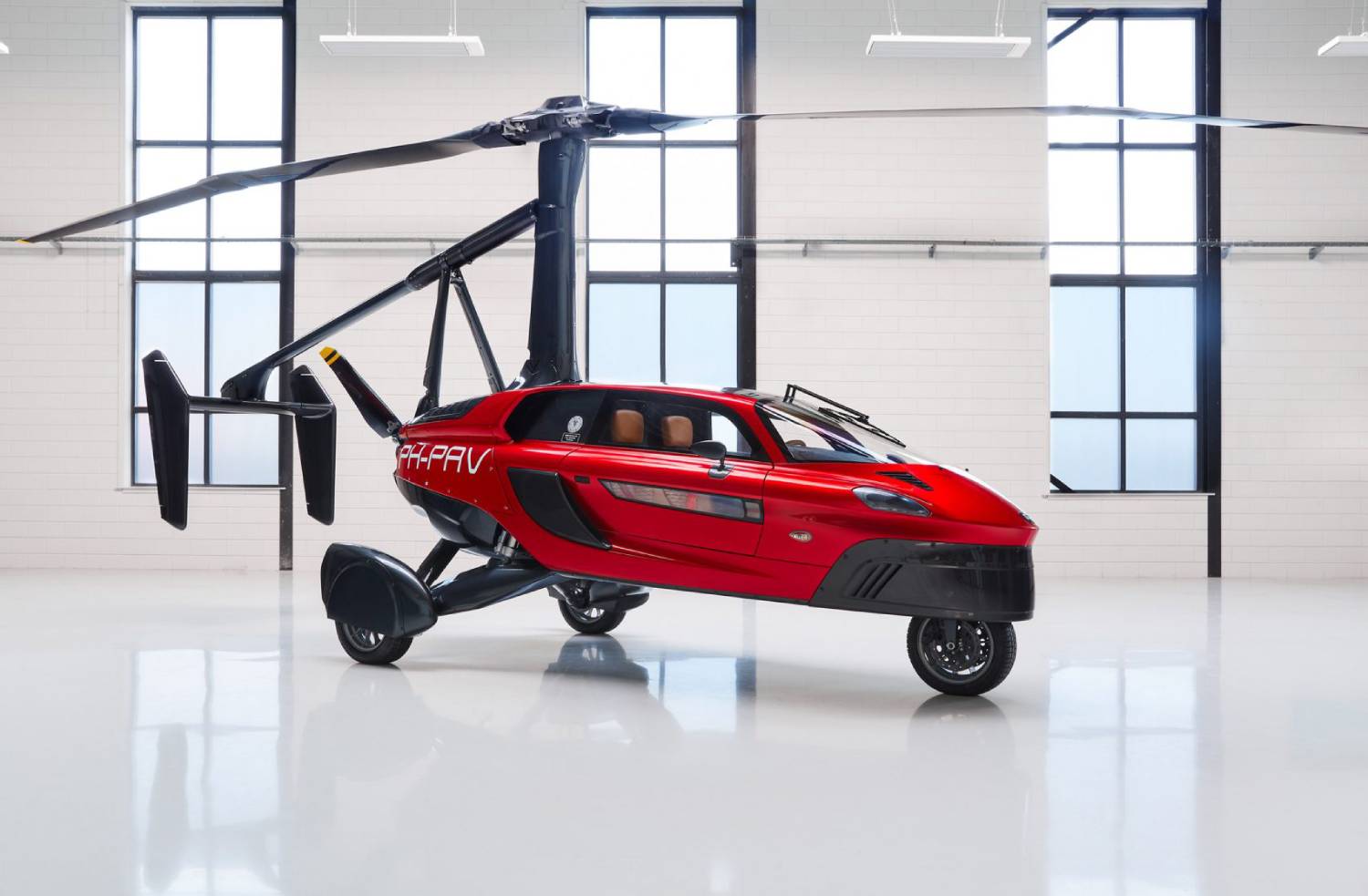 A red flying car with a helicopter rotor, showcasing its sleek design in a bright, spacious garage with large windows.