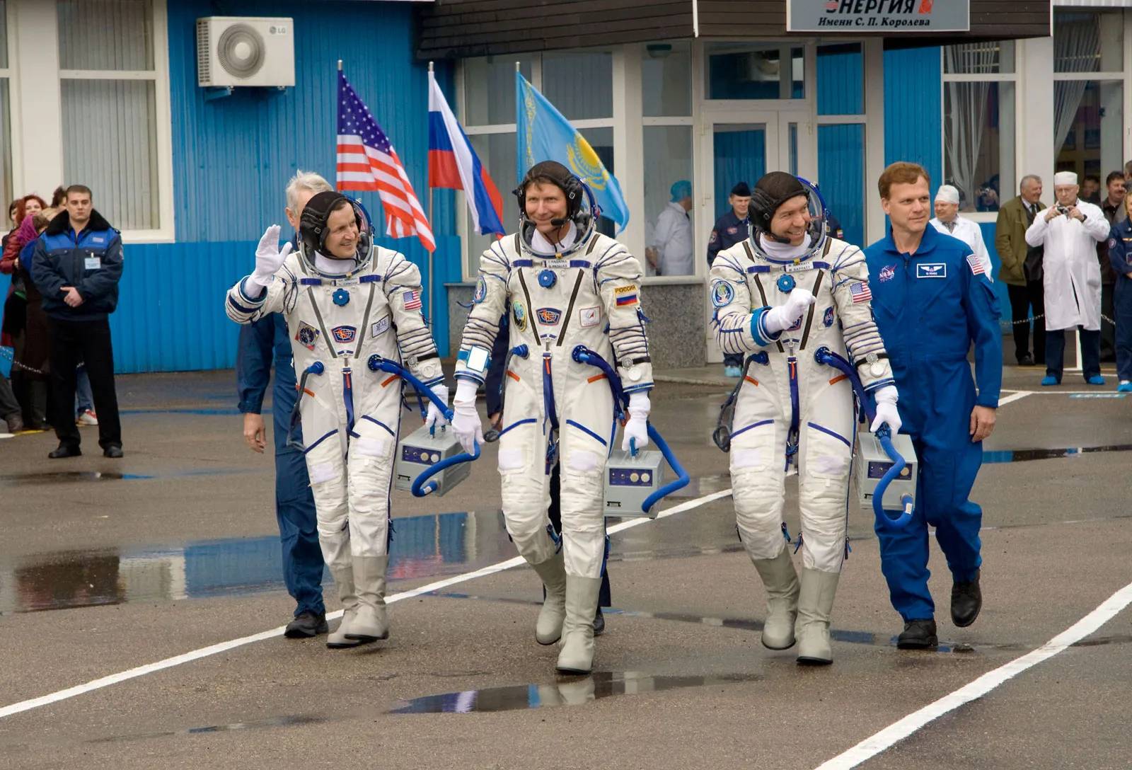 Astronauts in spacesuits walk together at a launch site, with flags of the USA, Russia, and Kazakhstan visible in the background.