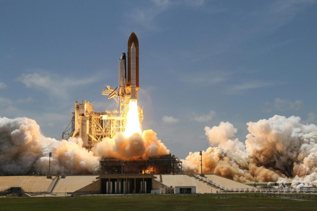 A space shuttle launches into the sky, flames and smoke billowing from its boosters against a clear blue backdrop.