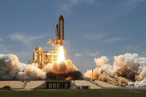 A space shuttle launches into the sky, flames and smoke billowing from its boosters against a clear blue backdrop.