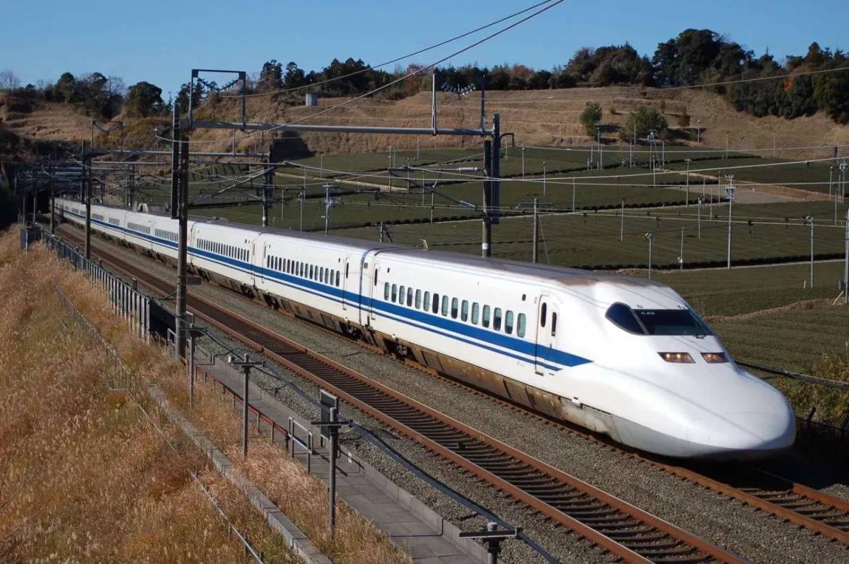 A sleek Shinkansen train speeds through green fields, showcasing its iconic white body with blue stripes against a clear blue sky.