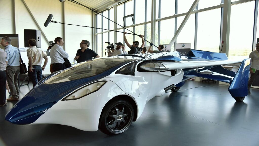 A futuristic flying car design showcased indoors, featuring sleek white and blue lines, drawing interest from onlookers and media.