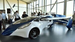 A futuristic flying car design showcased indoors, featuring sleek white and blue lines, drawing interest from onlookers and media.