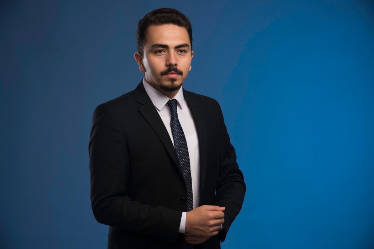  A man in a suit and tie stands confidently against a solid blue background.
 