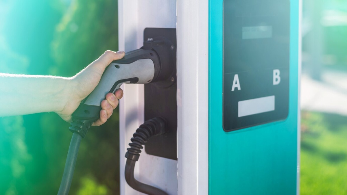    A person stands beside an electric car, holding the charging cable at an electric vehicle charging station.

