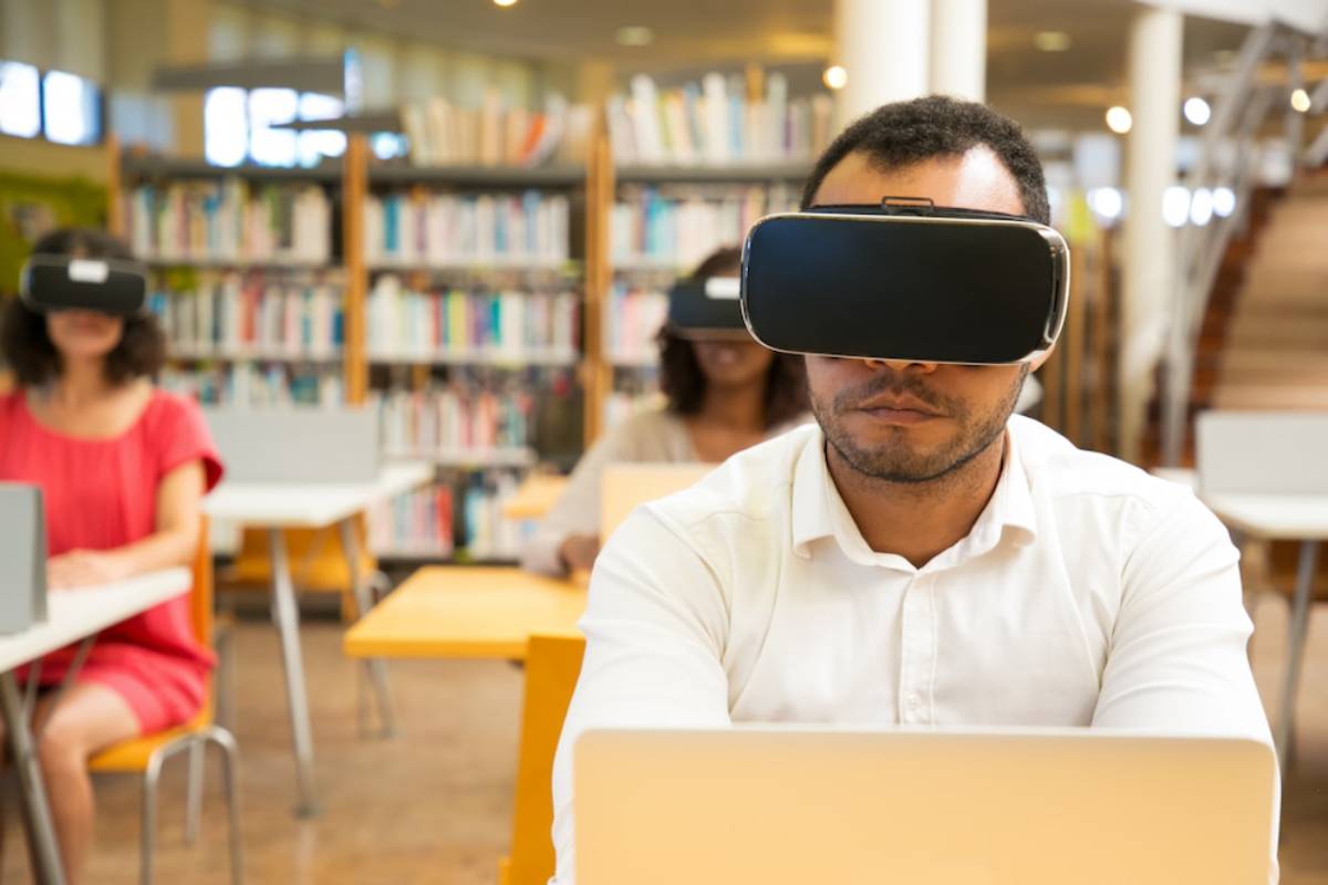  A man wearing virtual reality glasses stands in a library, surrounded by bookshelves filled with various books.
